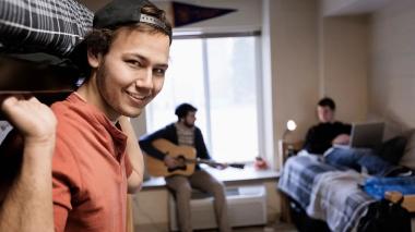 A young man in a light red shirt smiles toward the camera, leaning out from under a bunk bed. Behind him, two other young men play a guitar and work on a computer in this dorm room setting.