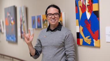 Salvador Sanchez stands in front of a wall featuring his art.  He holds up one hand, signing "Love you!" in ASL.