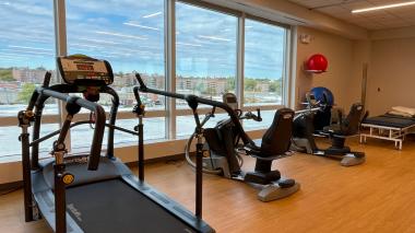 A row of rehabilitation and exercise machines along the walls of a  bright room with floor to ceiling windows and a hardwood style floor.