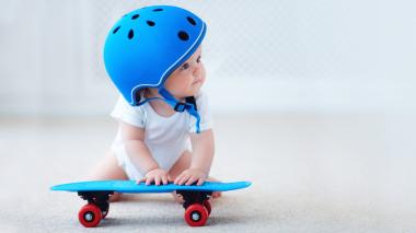 A baby sits on the floor wearing a helmet and playing with a small skateboard.