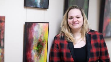 A smiling woman stands beside a wall full of framed artworks.