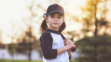 A girl in a baseball hat and little league shirt holds a bat on her shoulder.