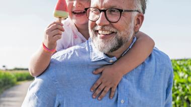 A happy man in a blue shirt carries a young child and their ice cream on his back.