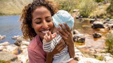 A woman hugs a baby close and smiles in a sunny outdoor scene with a lake in the background.