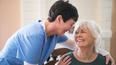 A caretaker and a seated patient smile at each other.