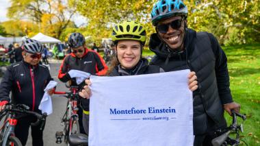 Two participants in the road race hold up a Montefiore Einstein towel from the road race. The're both dressed in bike helmets.