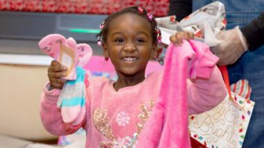 A young girl dressed in pink smiles widely, holding up presents.