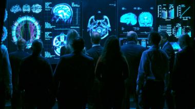 Doctors study a series of brain scans in a dark room. The scans glow blue on the screen.