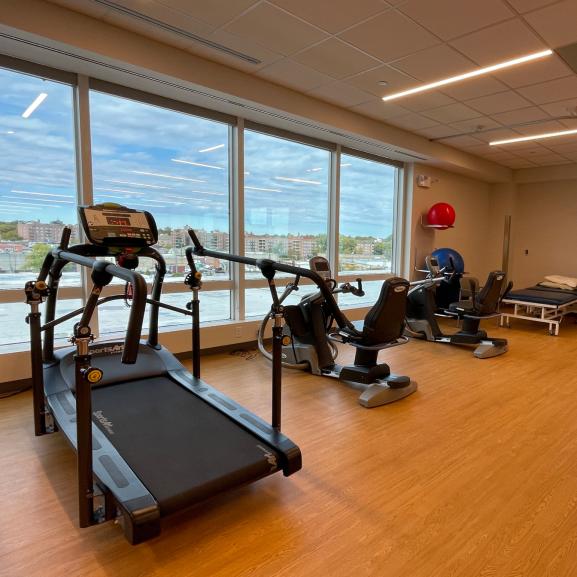 A row of rehabilitation and exercise machines along the walls of a  bright room with floor to ceiling windows and a hardwood style floor.